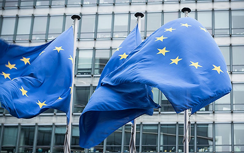 Three EU flags waving in the wind in front of glass buildings