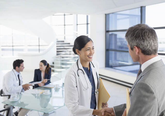A doctor shaking hands with a business professional while another doctor has a meeting in the background