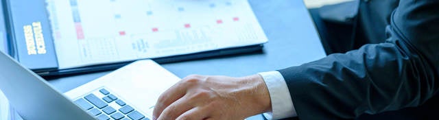 A person in a suit is taking notes and working on a laptop