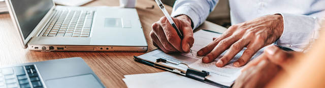 Person sitting at desk with a laptop taking notes on a clipboard