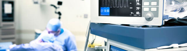 A closeup of operating room monitors while a surgeon works on a patient in the background