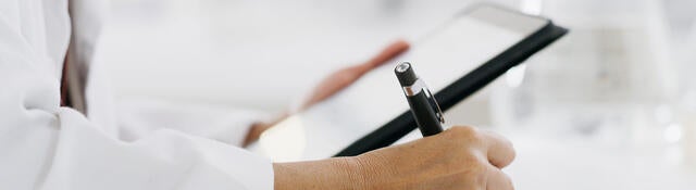 Closeup of a medical professional reading a tablet and making notes on paper while standing in a lab