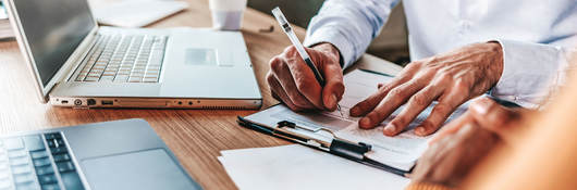 Person sitting at desk with a laptop taking notes on a clipboard