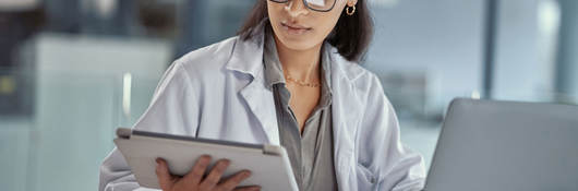 Doctor reading a tablet while working on a laptop