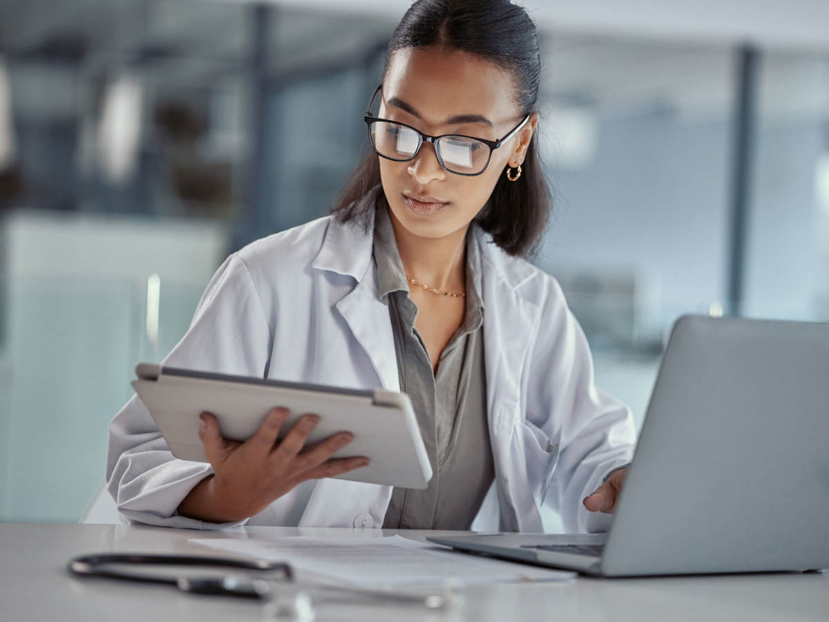 Doctor reading a tablet while working on a laptop