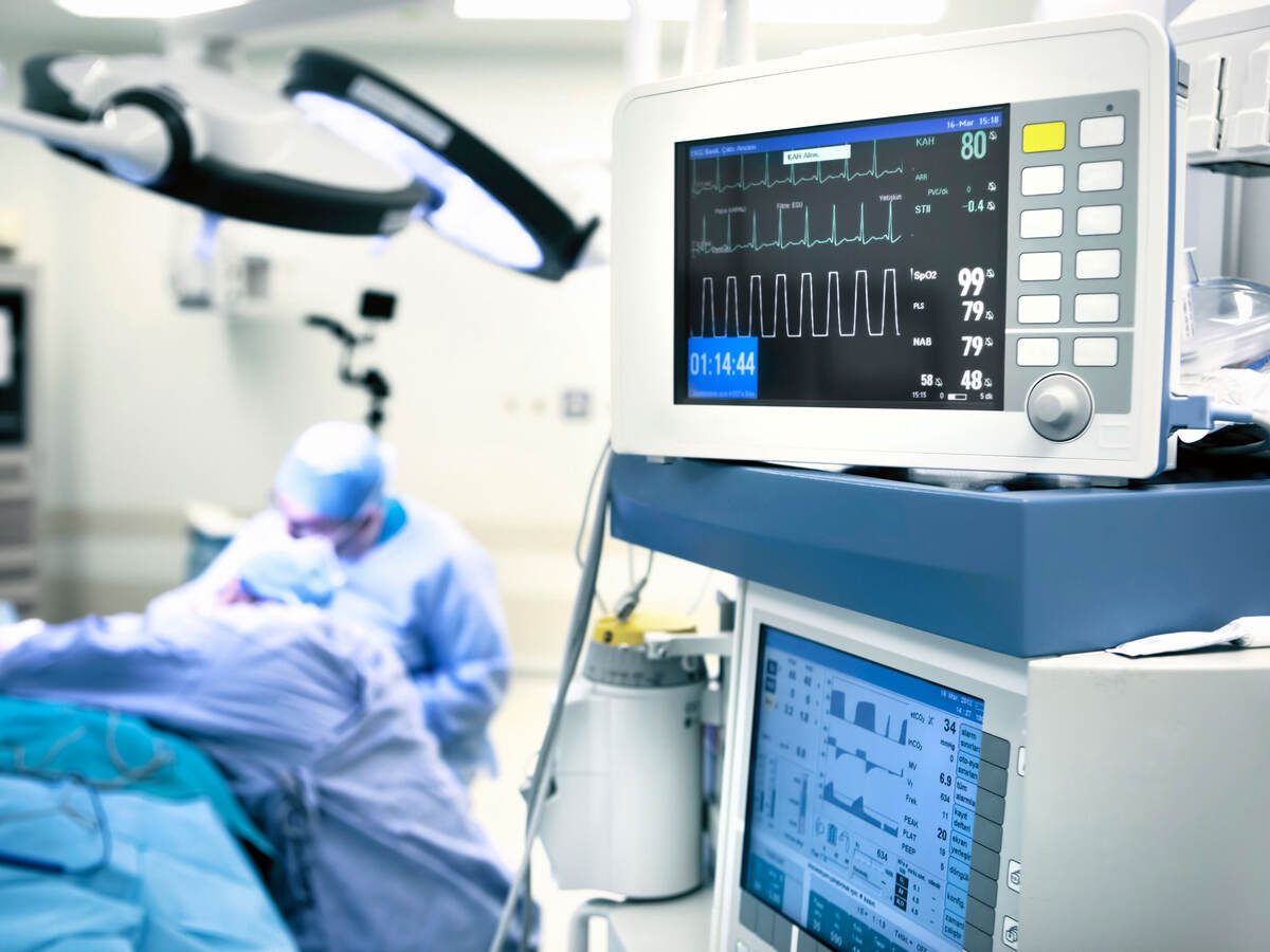 A closeup of operating room monitors while a surgeon works on a patient in the background